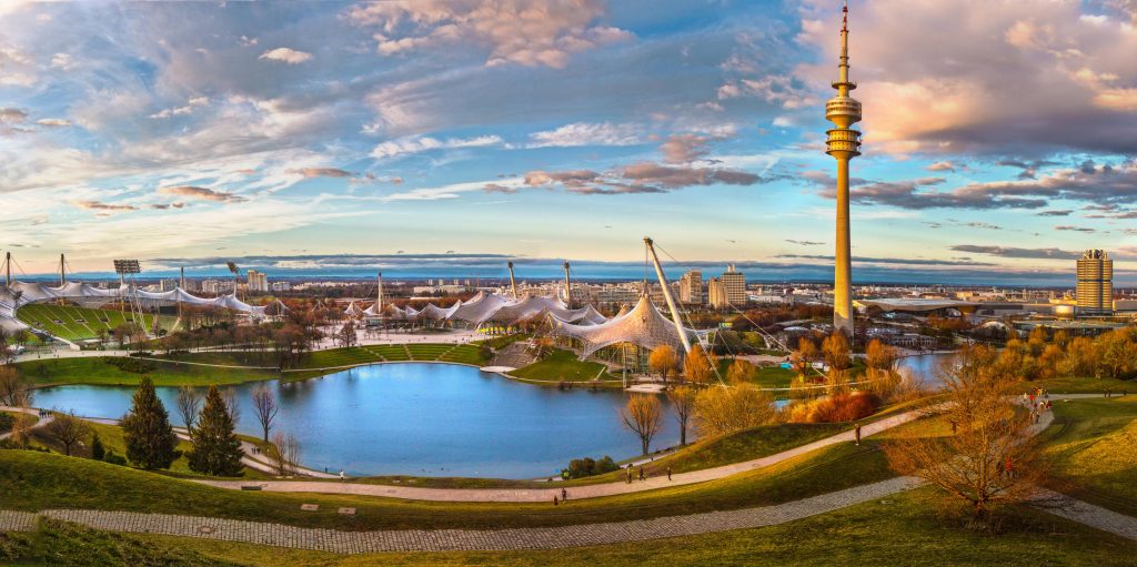 Panoramic view of Munich's Olympic Park at sunset, with the lake and iconic Olympic Tower in the foreground.