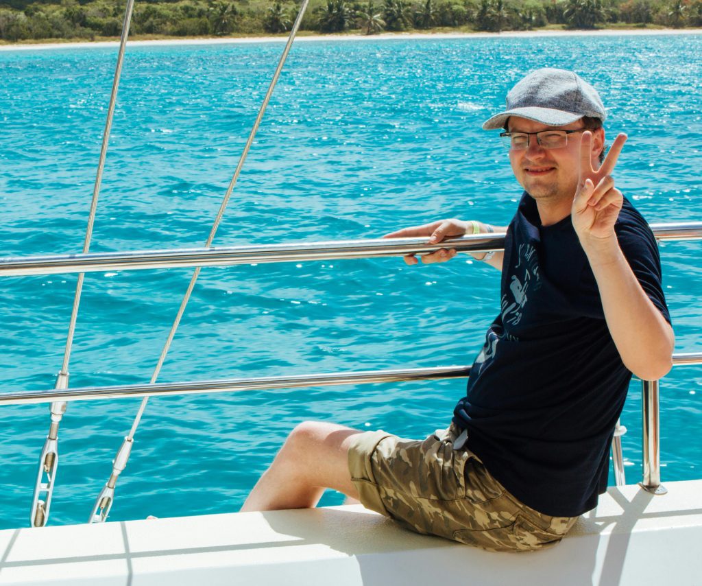 Caucasian man relaxing on a sailboat, posing with peace sign against the turquoise sea.