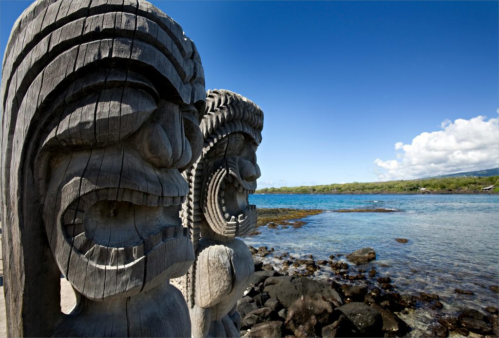 Wooden tiki statues by the ocean in Honaunau-Napoopoo, Hawaii. Bright day with rocky coastline.