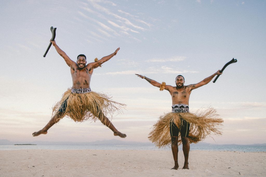 Two men in traditional attire joyfully jumping on a serene Fiji beach under a clear sky.