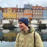 A young man in outerwear smiles at Nyhavn, Copenhagen, with colorful buildings in the background.