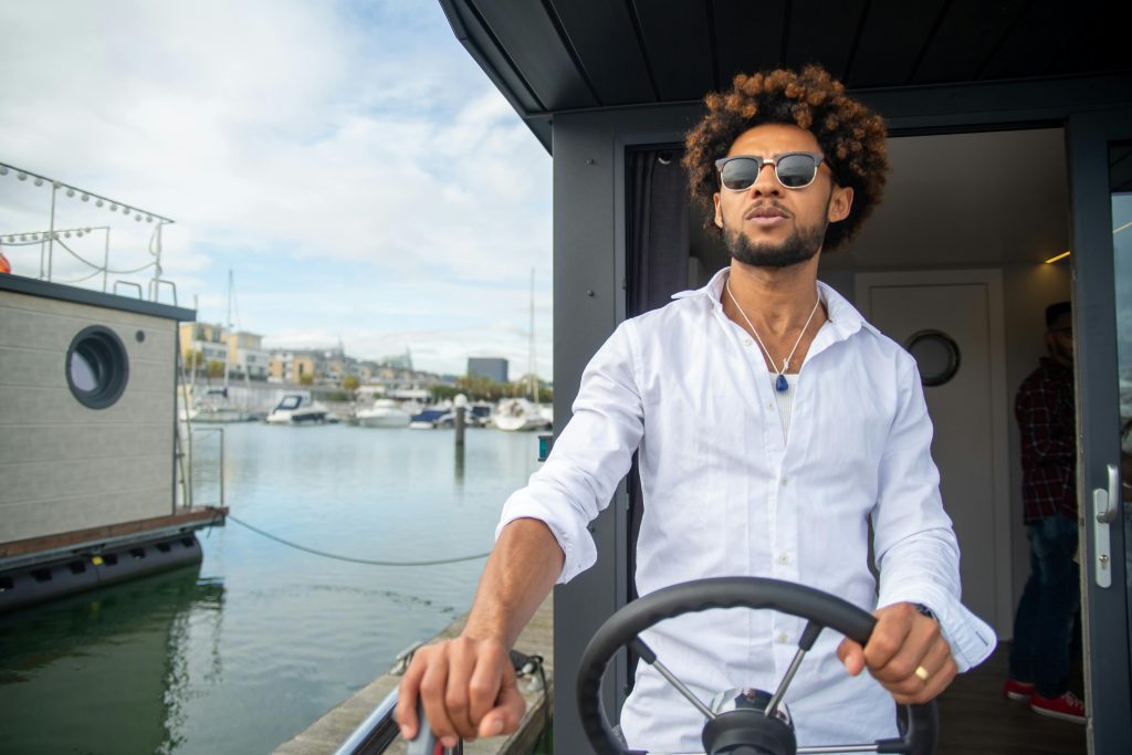 African American man with sunglasses steering a yacht in a sunny marina setting in Portugal.